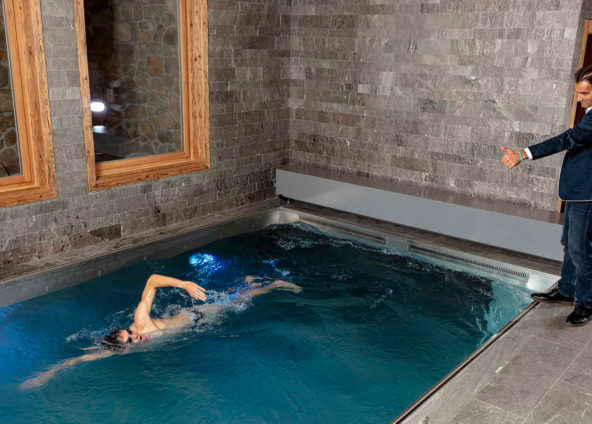 Ein Mann mit Sakko und Jeanshose steht am Beckenrand und hält seinen Daumen hoch. Er schaut auf einen Schwimmer welcher in dem Gegenstrompool von FlussPool trainiert.
