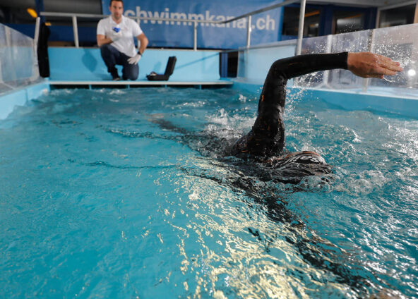 Ein ambitionierter Schwimmer in einem Schwimmanzug, schwimmt in einem Schwimmkanal in Horgen. Im Hintergrund ist ein Coach, welcher das Schwimmen beobachtet.